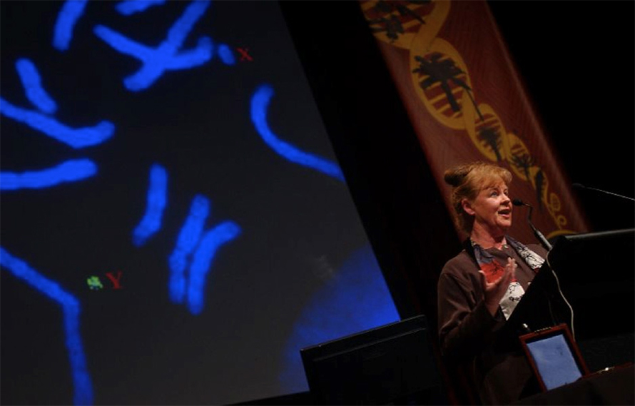 Graves gives a lecture. A slide of marsupial chromosomes is projected in the background.