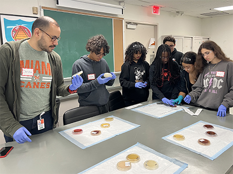Organizer/suspect Christian McDonald, at left, shows a group of Miami high school students how bacteria is linked to the crime scene and how various bacteria grow on agar plates.