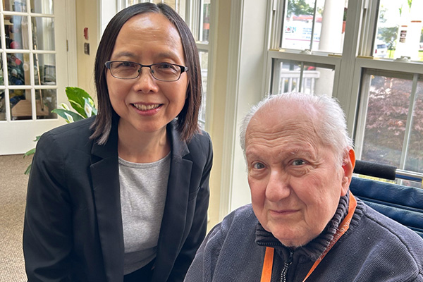 Guojuan (Amy) Liao stands beside Sanford Simon, seated in front of windows.