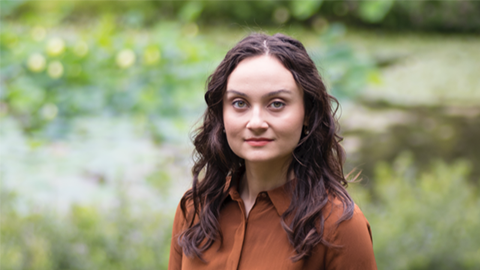 A portrait of Yuliya McAnany outdoors in a grassy area. She's wearing an orange blouse and has long, curly brown hair.